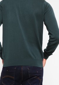 Mann in dunkelgrünem Pullover und dunkelblauen Jeans, von hinten gezeigt mit den Händen in den Hüften.