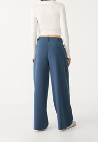 Femme portant un pantalon bleu large et un haut blanc moulant à manches longues, vue de dos sur un fond uni.