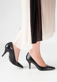 Escarpins en cuir verni noir à bout pointu et talon fin, portés avec une jupe plissée bicolore noire et crème.