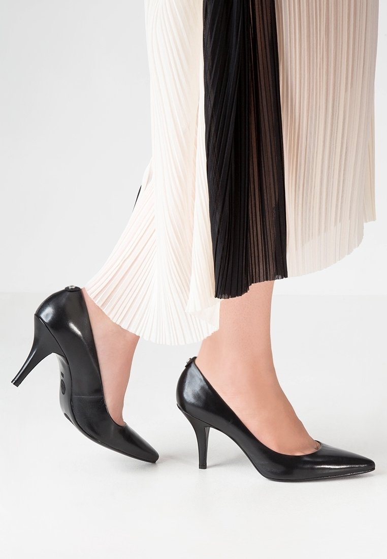 Escarpins en cuir verni noir à bout pointu et talon fin, portés avec une jupe plissée bicolore noire et crème.