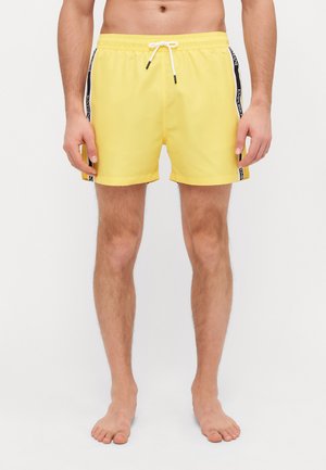Costumi da bagno gialli brillanti con vita elastica, cordoncino e strisce nere laterali con testo del marchio. Tessuto liscio e leggero.