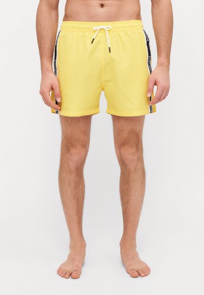Costumi da bagno gialli brillanti con vita elastica, cordoncino e strisce nere laterali con testo del marchio. Tessuto liscio e leggero.