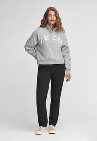 Grå sweatshirt med "POLO CLUB" tryckt i ljus färg, med en dragkedja vid kragen. Matchad med svarta byxor och ljusbeiga sneakers.