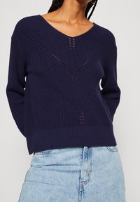 Navy blue knitted sweater with a V-neck, featuring a textured chevron pattern and open knit details, paired with light blue denim.