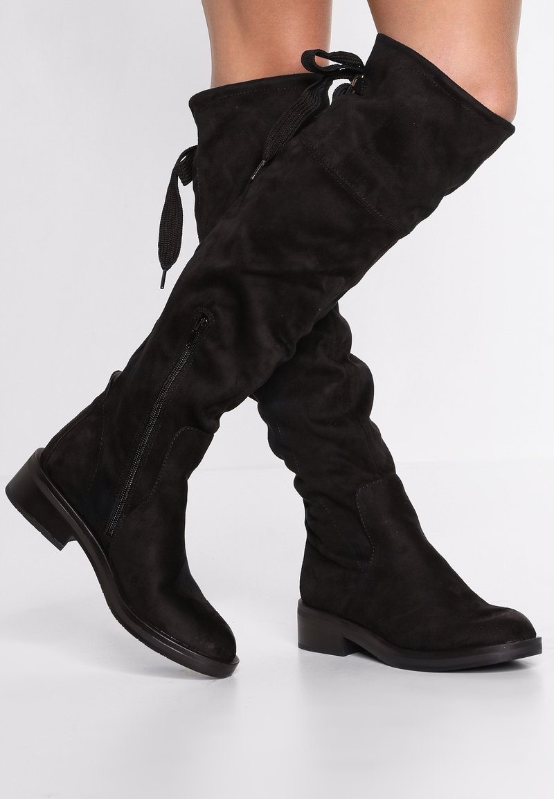 Bottes noire en daim montantes au-dessus du genou avec talons blocs bas, fermetures éclair latérales et liens en lacets en haut, portées sur des jambes croisées.