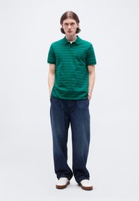 Homme debout les mains dans les poches, portant un polo rayé vert, un jean ample foncé et des baskets blanches, sur fond uni.