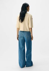 Pull en beige cropped et jean bleu à jambes larges. Le pull a des manches courtes ; le jean est en denim délavé avec des poches arrière. Fond propre.