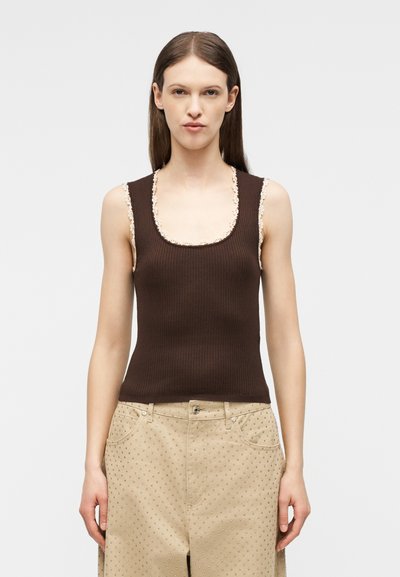 Γυναίκα που φοράει ένα καφέ αμάνικο ριπ τοπ με δαντέλα και μπεζ παντελόνι με διακριτικό πουά σχέδιο, στέκεται μπροστά σε απλό φόντο.