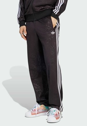 Person trägt schwarze Adidas-Trainingshose mit weißen Streifen und eine schwarze Jacke, Hände teilweise in den Taschen, bunte Adidas-Sneaker sichtbar.