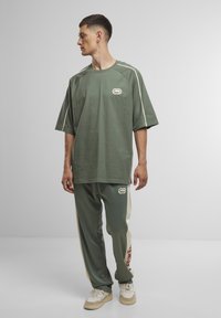 Oversized groene katoenen t-shirt met witte accenten, gecombineerd met bijpassende groene joggingbroek met zijstrepen en rode letters.