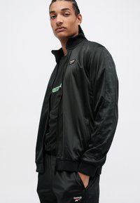 Jaunas vyras su pinto plaukais, vilkintis juodą Reebok treniruotę su logotipais striukės krūtinėje, rankovėse ir kelnių kišenėje, stovi su rankomis kišenėse.