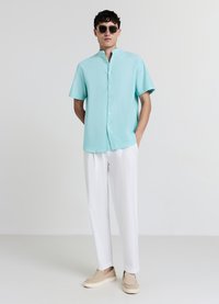 Hellblaues Kurzarmhemd mit Stehkragen, weiße Hose und beigen Slip-On-Schuhen, stehend vor einfachem Hintergrund.