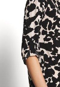 Svart och beige blusa med ett abstrakt djurmönster, rullade ärmar med knappdetaljer och en mjuk, lätt tygkvalitet.