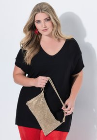 Černé tričko s výstřihem do V a krátkými rukávy, spárované s červenými texturovanými kalhotami; drží zlatou metalickou kabelku s řetízkovým popruhem.