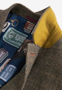 Grijze geruite wollen jas met een gele vilten kraag. De binnenkant heeft een blauwe voering met ontwerpstukken van tickets en andere grafische ontwerpen.