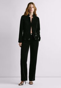 Naine kannab avatud mustast sametist jopet ja sobivaid laia säärega pükse, seisab helelilmelise tausta ees mustade rihmikandadega kontsakingades.