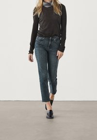 Woman wearing a black long-sleeve top, dark blue cropped jeans, black loafers, and a plaid neck scarf, walking on a gray floor.