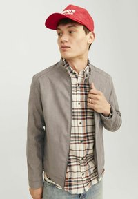 Jachetă din suede gri cu guler ridicat peste o cămașă în carouri în nuanțe de crem, albastru marin și roșu. Modelul poartă o șapcă roșie de baseball. Fundal simplu.