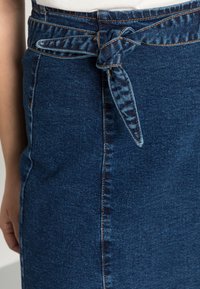 Saia de ganga com uma cintura cingida por um cinto, apresentando uma cor azul escuro, costura azul clara e uma textura suave. Inclui um detalhe de nó no cinto.