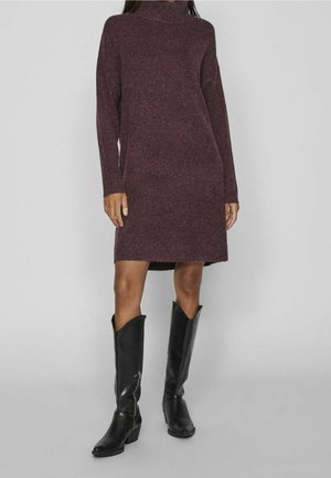 Žena v dlouhých rukávech tmavě fialových pletených šatech a černých kožených kozačkách nad kolena před jednobarevným šedým pozadím.