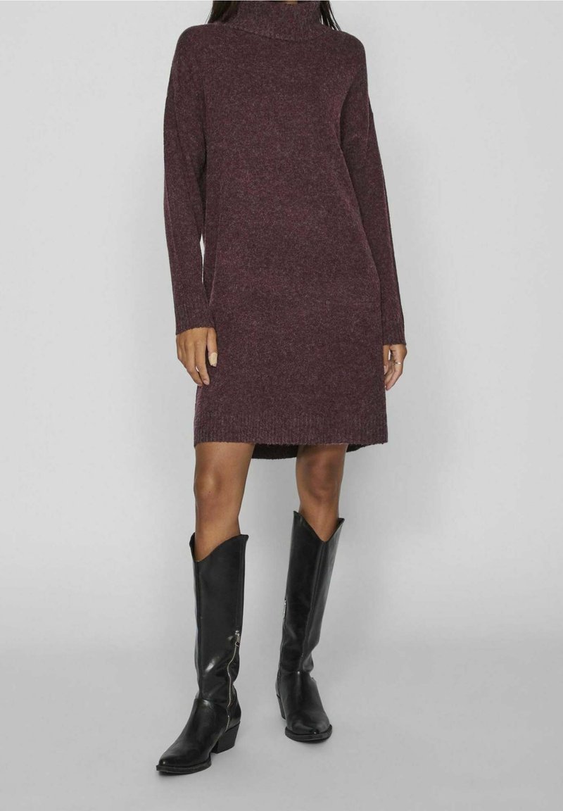 Femme portant une robe en maille violette foncée à manches longues et des bottes en cuir noires montant jusqu'au genou, sur un fond gris uni.