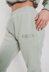 Hellgrüne Sweatpants aus Baumwollmischung, mit elastischem Bund und Seitentaschen. "CALUA" in hellerer Farbe auf dem Oberschenkel gestickt.