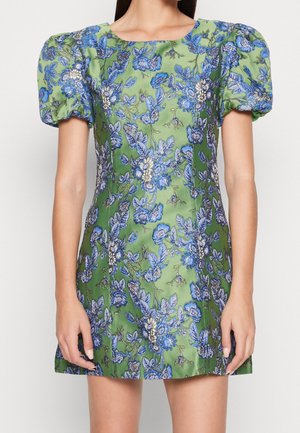 Femme portant une mini robe verte à imprimé floral bleu et manches courtes bouffantes, debout les mains détendues le long du corps.