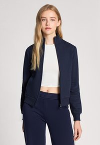 Felpa blu navy con zip, realizzata in tessuto morbido, con colletto alto e polsini a coste. Indossata con un top bianco e pantaloni blu navy coordinati.