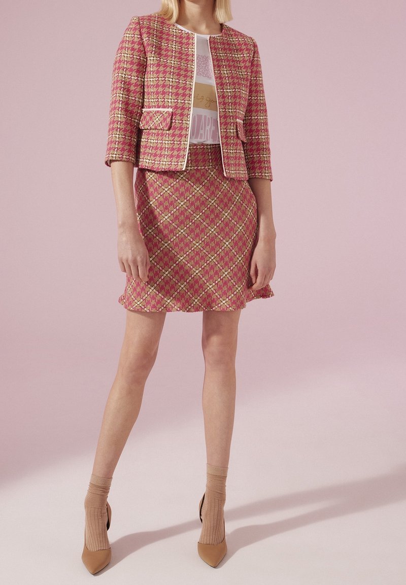 Vrouw die een roze en geel geruit rokpak draagt met driekwartmouwen, gecombineerd met een wit shirt met print en beige hakken met sokken.