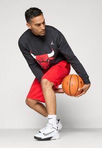 Melnais sporta krekls ar sarkano bulli, sarkanas šortus un baltas Nike kedas; tur rokās oranžs basketbola bumba, demonstrējot sporta apģērbu.