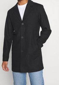 Abrigo largo negro con cuello en pico, cierre de un solo botón en la parte delantera, tres botones y bolsillos laterales, combinado con una camiseta blanca y jeans azules.