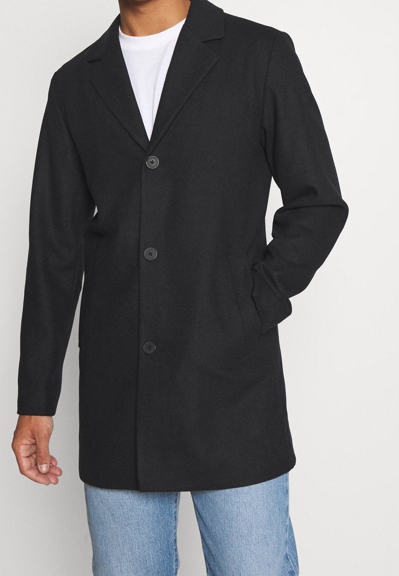 Abrigo largo negro con cuello en pico, cierre de un solo botón en la parte delantera, tres botones y bolsillos laterales, combinado con una camiseta blanca y jeans azules.