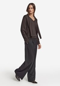 Mörkbrun v-ringad tröja med huva i samma färg, kombinerad med löst sittande svarta byxor och svart spetsiga skor. Slät textur.