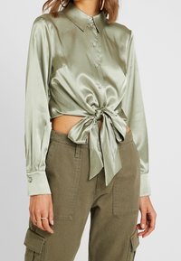 Kvinna iförd en ljusgrön satinblus med lång ärm, knäppt framtill och knuten i midjan, tillsammans med olivgröna cargobyxor och guldörhängen. 