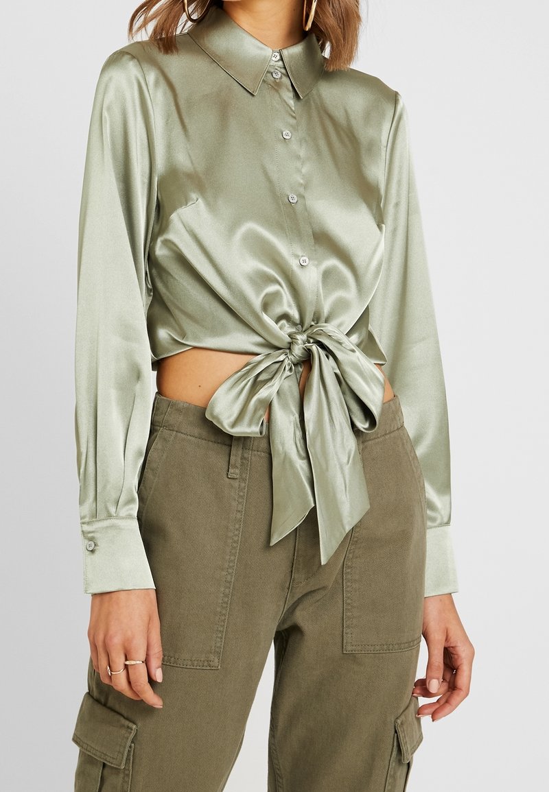 Kvinna iförd en ljusgrön satinblus med lång ärm, knäppt framtill och knuten i midjan, tillsammans med olivgröna cargobyxor och guldörhängen. 