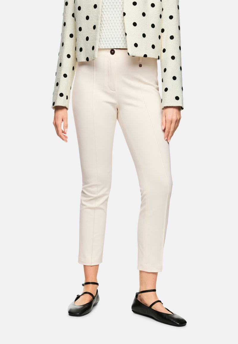 Helle rosa maßgeschneiderte Hose mit Vordernähten, kombiniert mit einem strukturierten weißen Oberteil und einer gepunkteten Jacke, ergänzt durch schwarze flache Schuhe.