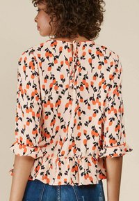 Blouse pêche avec motif floral noir et orange, manches 3/4, ourlet festonné et fermeture au dos avec lien. Tissu lisse avec une légère texture.
