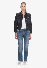 Veste en denim noir avec des boutons sur le devant, deux poches poitrine, débardeur blanc, jeans en denim bleu et baskets clair avec des lacets gris.