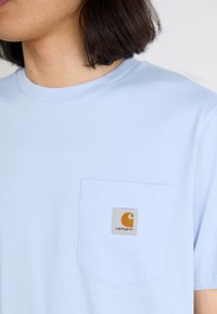 Light blue cotton t-shirt with a round neckline, short sleeves, and a chest pocket featuring a yellow and grey Carhartt logo patch.