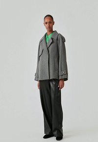 Woman wearing a grey oversized coat over a green shirt and wide black leather trousers, standing against a plain light background.