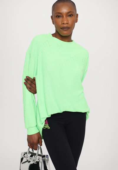 Felpa oversized verde neon con tessuto texture, spalle scese e scollatura rotonda, abbinata a leggings neri con accenti colorati.