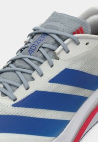 Άσπρο αθλητικό παπούτσι με μπλε διαγώνιες ρίγες. Διαθέτει γκρι κορδόνια και υφασμένη επάνω επιφάνεια. Το λογότυπο είναι ορατό στο πλάι και στη γλώσσα του παπουτσιού.