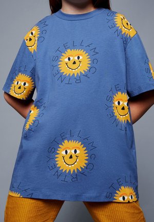 T-shirt en coton bleu avec des motifs de soleil jaunes répétitifs souriants et du texte noir autour du soleil, manches courtes et coupe décontractée.