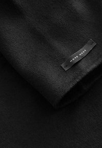 Tissu en laine noir plié avec une étiquette noire cousue portant la mention "HAND MADE" en texte blanc.