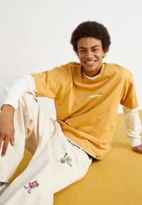 Tricou cu mânecă scurtă de culoare muștar, cu logo alb, suprapus peste un cămașă albă cu mânecă lungă; pantaloni crem cu desene colorate brodate, reprezentând o floare și un desen animat.