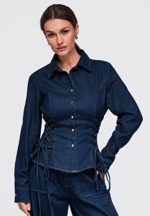 Femme portant une veste en jean bleu foncé avec des côtés lacés et un pantalon en jean assorti, debout avec une main sur la hanche.