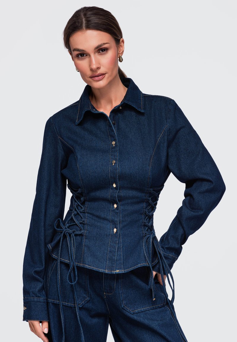 Femme portant une veste en jean bleu foncé avec des côtés lacés et un pantalon en jean assorti, debout avec une main sur la hanche.