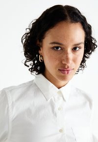 Camisa blanca de botones con cuello, que presenta una tela texturizada y un bolsillo en el pecho. El modelo tiene el cabello rizado y aretes de aro dorados.