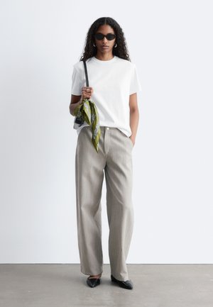 Femme portant des lunettes de soleil noires, un t-shirt blanc, un pantalon large beige, des chaussures noires, tenant un sac noir avec une écharpe verte, debout sur un sol gris.