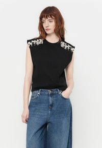 Svart ermeløs topp med pyntede skulderdetaljer, laget av et mykt stoff, kombinert med høy midje blå jeans.